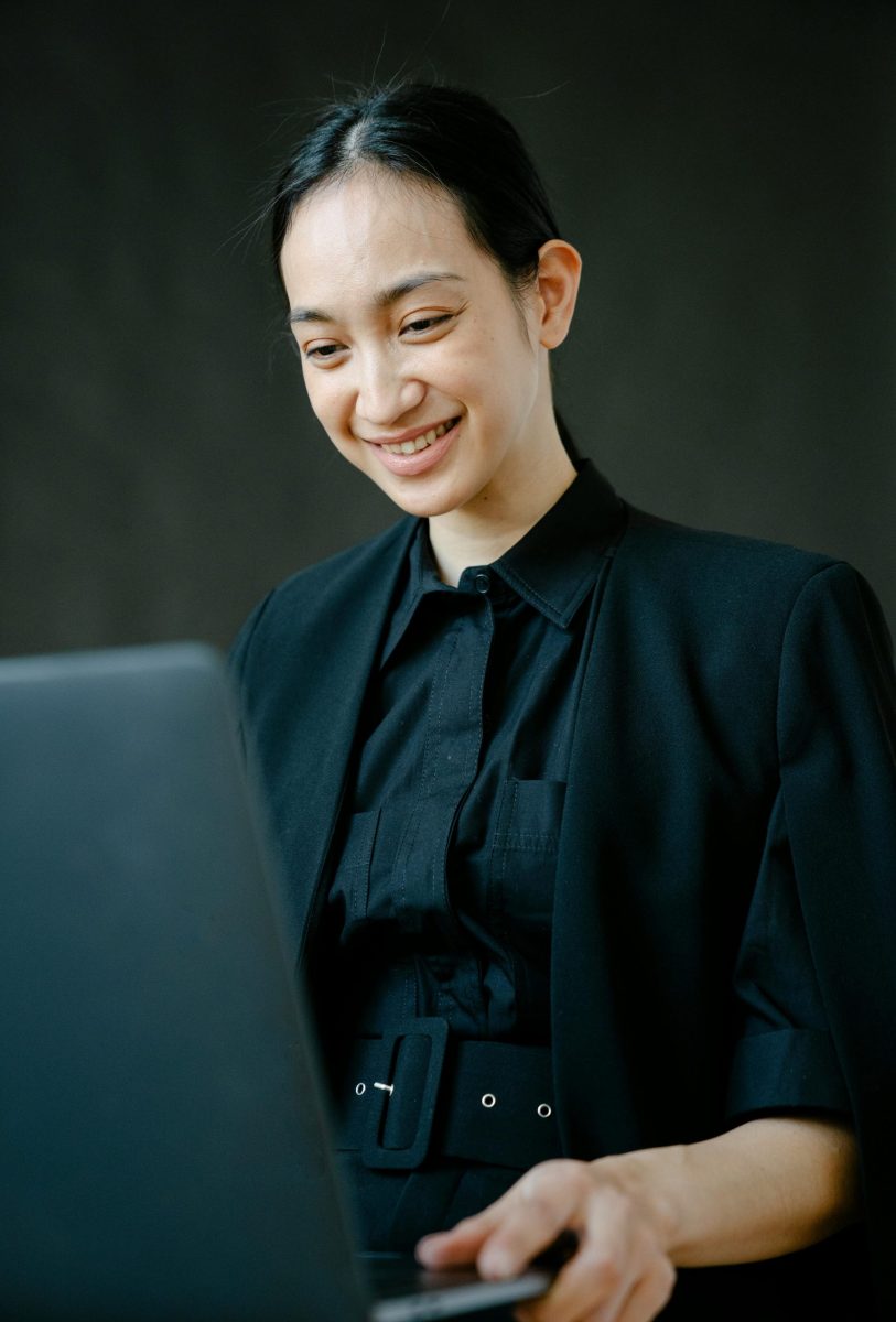 Low angle of positive ethnic female entrepreneur in formal wear using netbook against black background