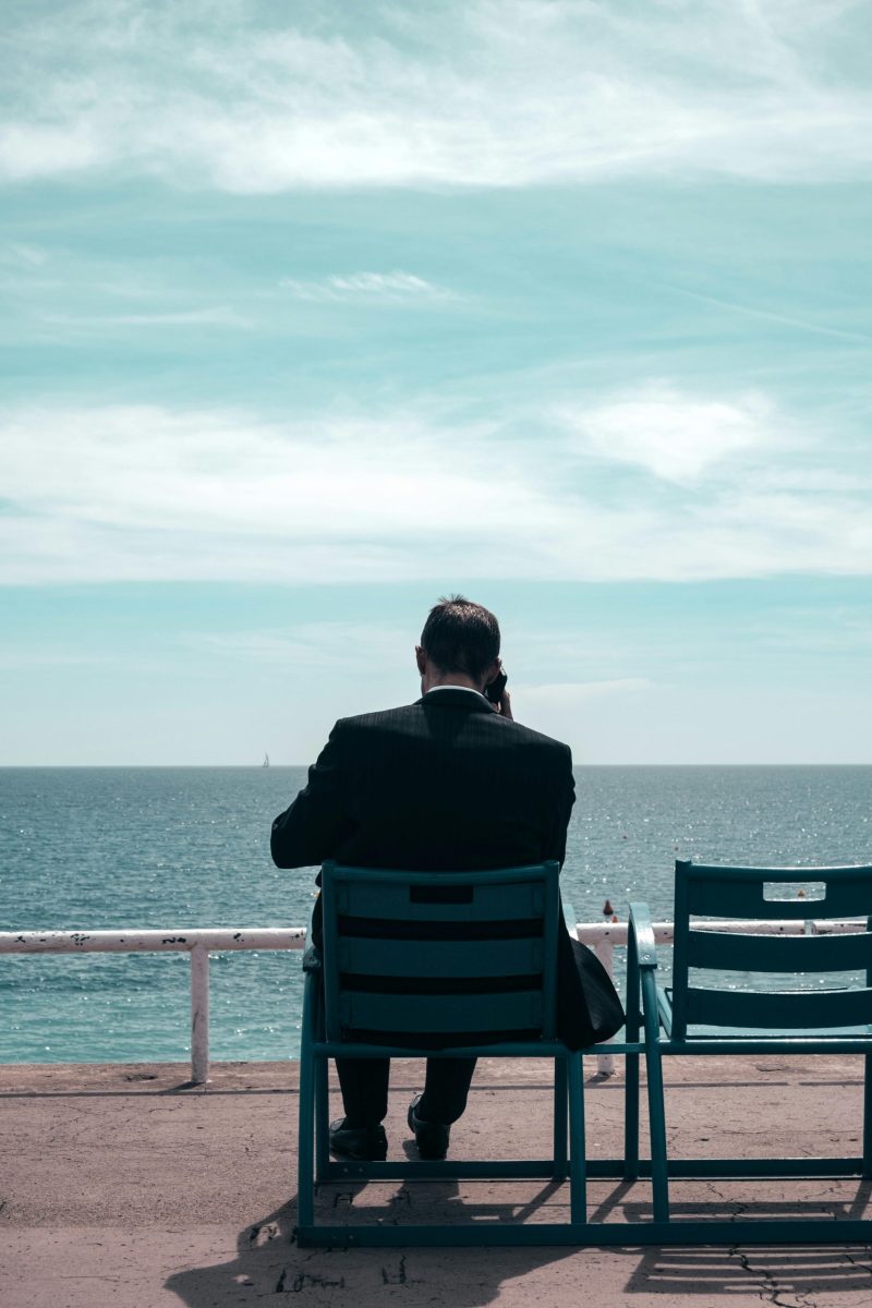 A man in a suit sits on a bench, gazing at the sea on a sunny day.